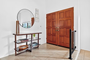 A hallway with a wooden door, a mirror, a console table with books and decorative items, and a tiled floor. - 3-Bedroom Desert Oasis Just Steps to La Quinta Resort! (La Quinta)