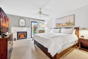 A bedroom with a bed, a fireplace, a ceiling fan, a TV, a desk, and a window with a view of a garden. - 3-Bedroom Desert Oasis Just Steps to La Quinta Resort! (La Quinta)