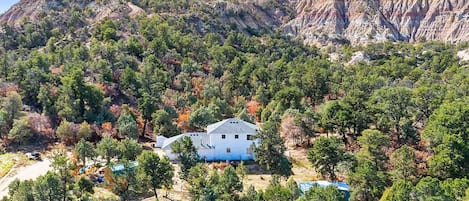 Un edificio blanco con tejado a dos aguas rodeado de árboles frondosos y montañas al fondo.