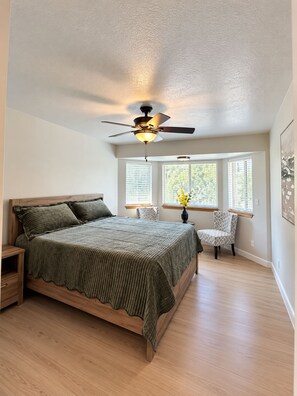 A bedroom with a bed, a chair, a nightstand, a ceiling fan, and a window with blinds.