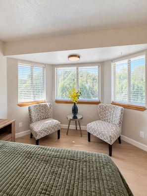 A room with a bed, two patterned chairs, a small table with a vase, and large windows with blinds.