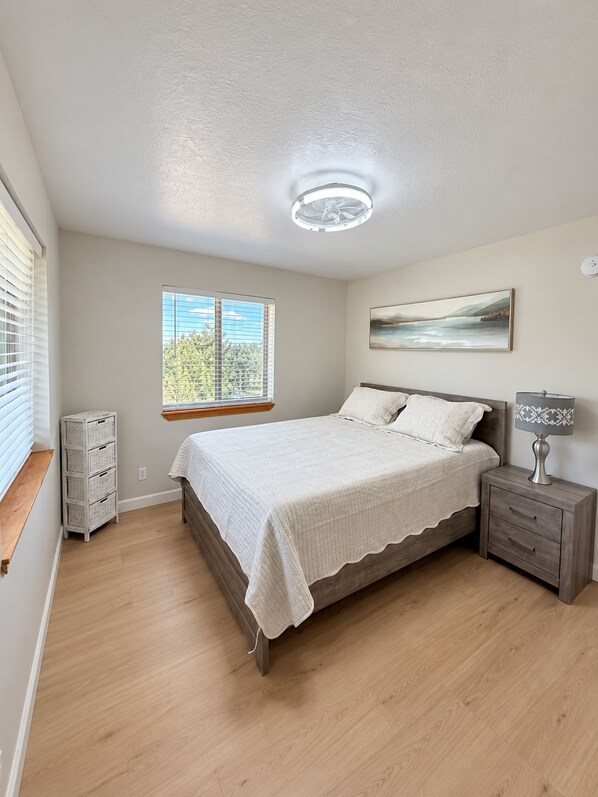 A bedroom with a bed, bedside table, lamp, window, and artwork on the wall.