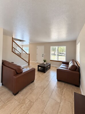 A spacious living room with two brown leather sofas, a wooden staircase, and a white door.
