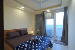A hotel room with a bed, a nightstand, a lamp, a ceiling fan, and a sliding glass door leading to a balcony. - Forest view 1BHK Premium studio appartment (New Delhi)