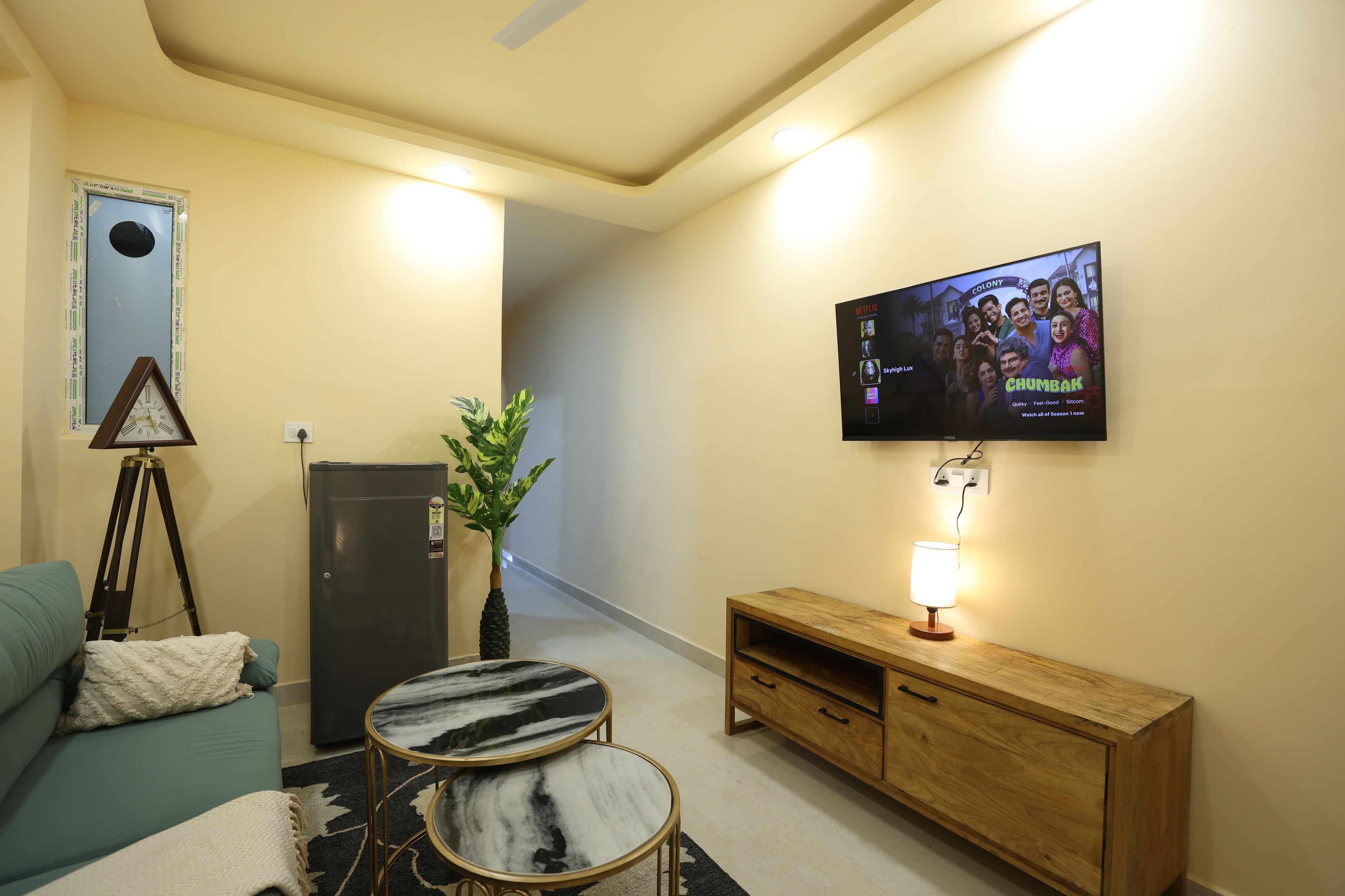 Una sala de estar con televisión montada en la pared, una mesita, un sofá, un armario y una planta.