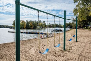 A sandy beach with a swing set and a view of a lake with boats. - Cozy Corner at Sommer Dorf (West Branch)