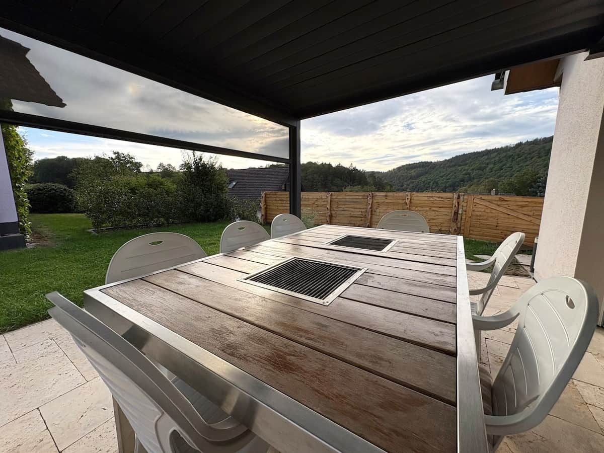 Une terrasse extérieure avec une table et des chaises en bois, un espace abrité et une vue sur un paysage verdoyant.