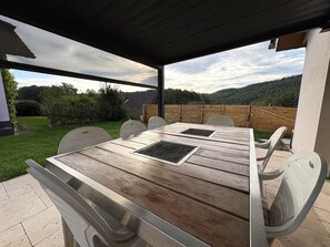Une terrasse extérieure avec une table et des chaises en bois, un espace abrité et une vue sur un paysage verdoyant.