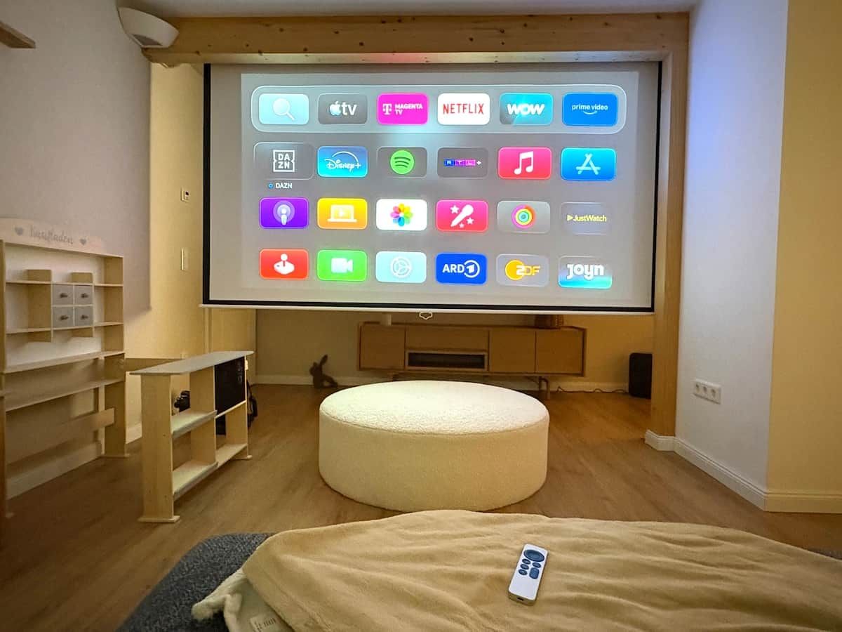 A room with a large screen displaying various streaming apps, a round ottoman, and a bed with a remote control.