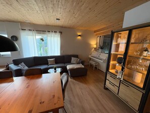 A modern living room with a wooden ceiling, a dark sofa, a dining table, and a glass cabinet with wine glasses.