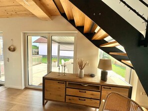 Une pièce avec un bureau en bois, une lampe et un vase contenant des plantes séchées. Une grande fenêtre offre une vue sur une maison et une pelouse.