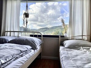 A bedroom with two beds, a window with a view of a forested landscape, and a hanging plant.