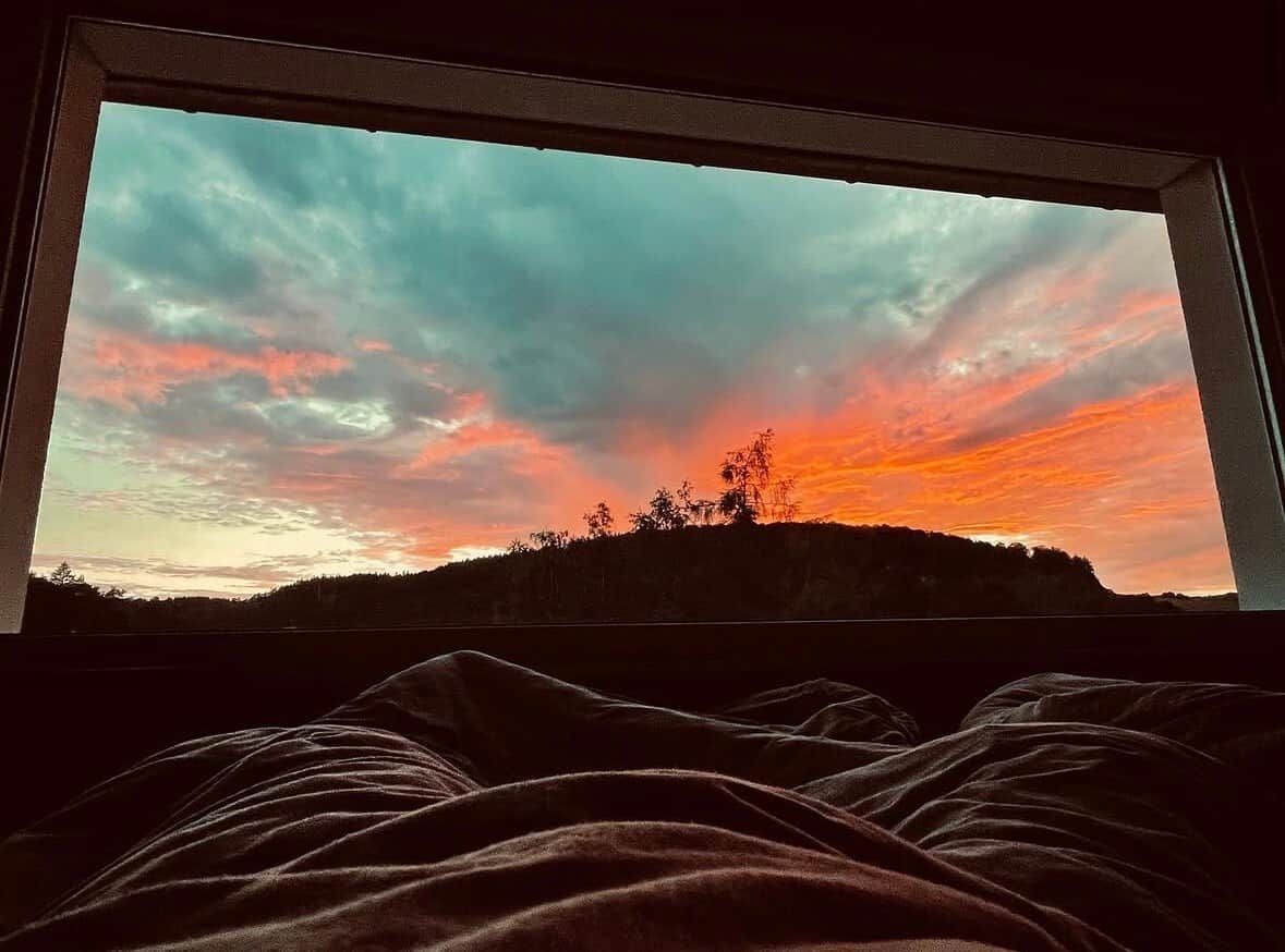 A window with a view of a sunset over a hill, with a blanket partially visible in the foreground.