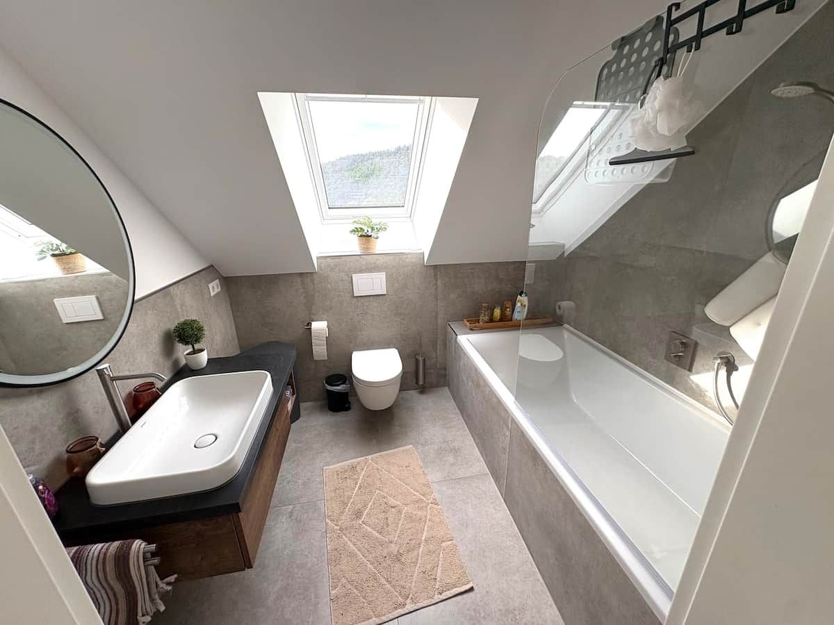 Ein modernes Badezimmer mit einer freistehenden Badewanne, einem Waschbecken und einer Toilette. Über der Toilette befindet sich ein Oberlicht und ein kleiner Topfpflanze auf dem Fensterbrett.