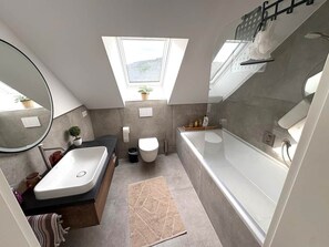 Une salle de bain moderne avec une baignoire sur pieds, un lavabo et des toilettes. Une lucarne se trouve au-dessus des toilettes et une petite plante en pot est posée sur le rebord de la fenêtre.