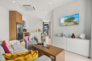 A modern living room with a sofa, coffee table, and a dining area with a white cabinet and a painting of a beach scene. - 1BR Retreat | Pool & Jacuzzi | Near Eagle Beach (Noord)