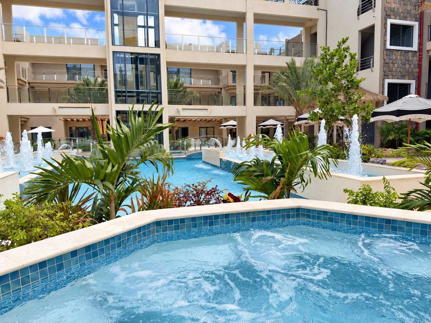 A hotel pool area with multiple levels, water fountains, and palm trees.