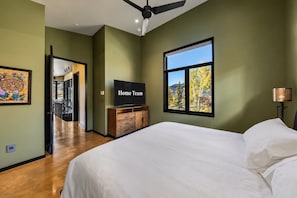 Ein Schlafzimmer mit einem Bett, einem Fernseher auf einer Holzkommode, einem Fenster mit Blick auf Bäume und einem Deckenventilator.