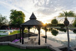 A gazebo with a tiled roof, a small table, and benches, surrounded by a water feature and palm trees. - Lakin' It Easy | Luxe Waterfront Oasis | Sleeps 16 (Mesa)