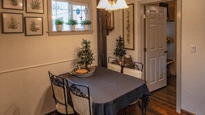 Un comedor con una mesa dispuesta para dos, sillas, un pequeño árbol de Navidad y una ventana con plantas en macetas.