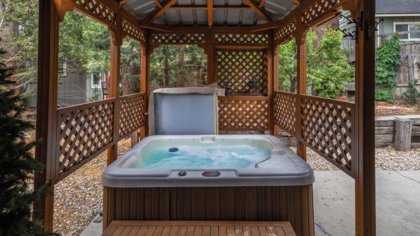 Un jacuzzi en un gazebo de madera con diseño de celosía, rodeado de árboles y una valla de madera.