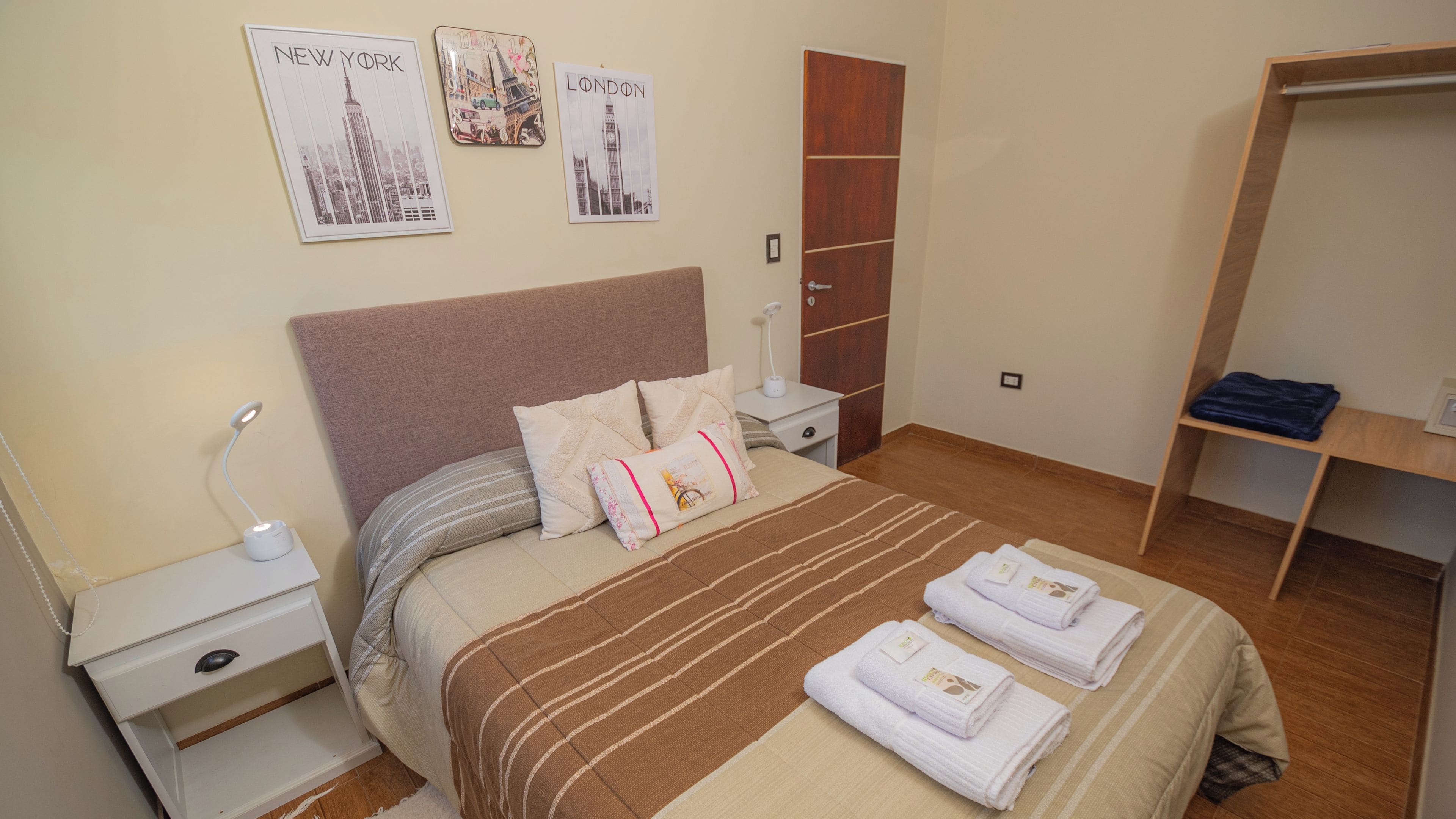 Ein ordentlich eingerichtetes Schlafzimmer mit einem Bett, Nachttischen, einem Kleiderschrank und Wandbildern bekannter Wahrzeichen.