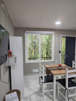Una habitación con una mesa de comedor, sillas, una ventana que da a árboles, una puerta blanca y una pared blanca.
