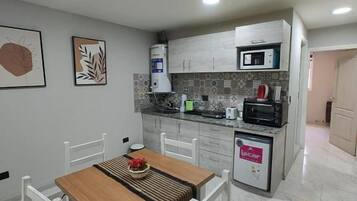 Una cocina compacta con armarios blancos, microondas y cafetera. Hay una mesa de comedor con sillas y una pequeña alfombra.