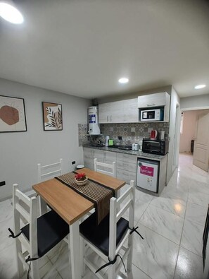 Una cocina compacta con armarios blancos, microondas y cafetera. Hay una mesa de comedor con sillas y una pequeña alfombra.