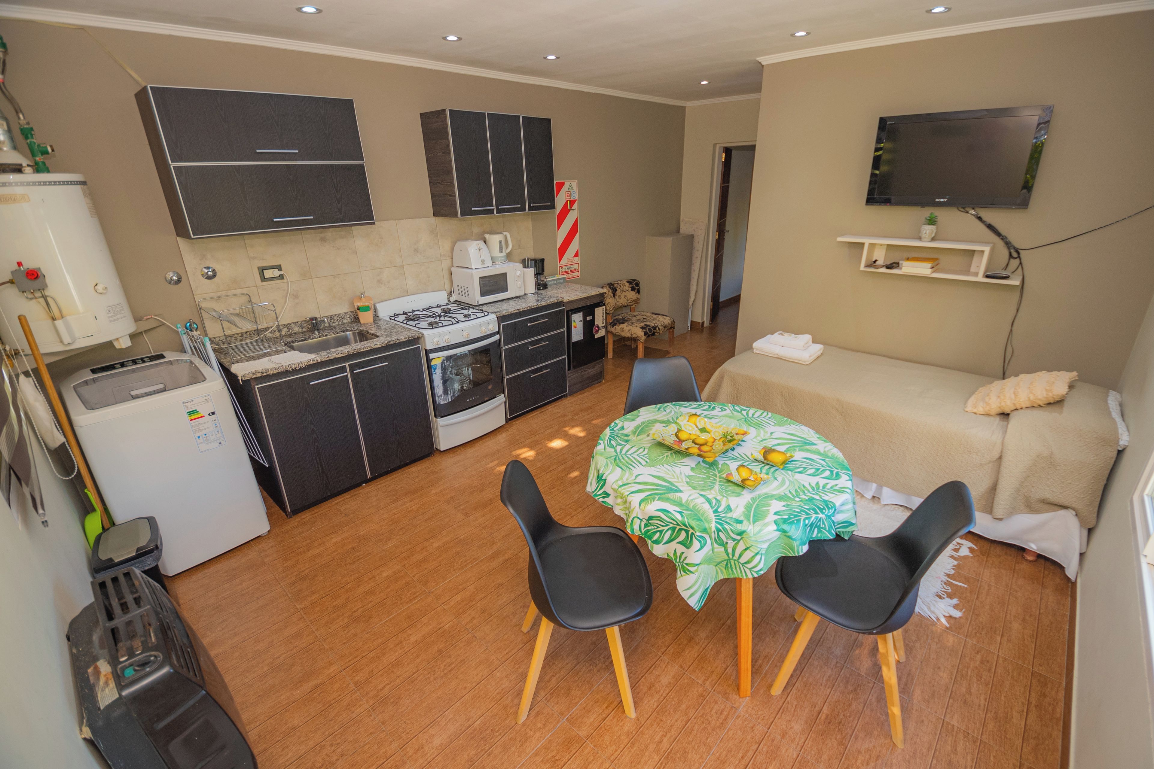 A compact living space with a kitchenette, a dining area with a table and chairs, a sofa, and a flat-screen TV mounted on the wall.