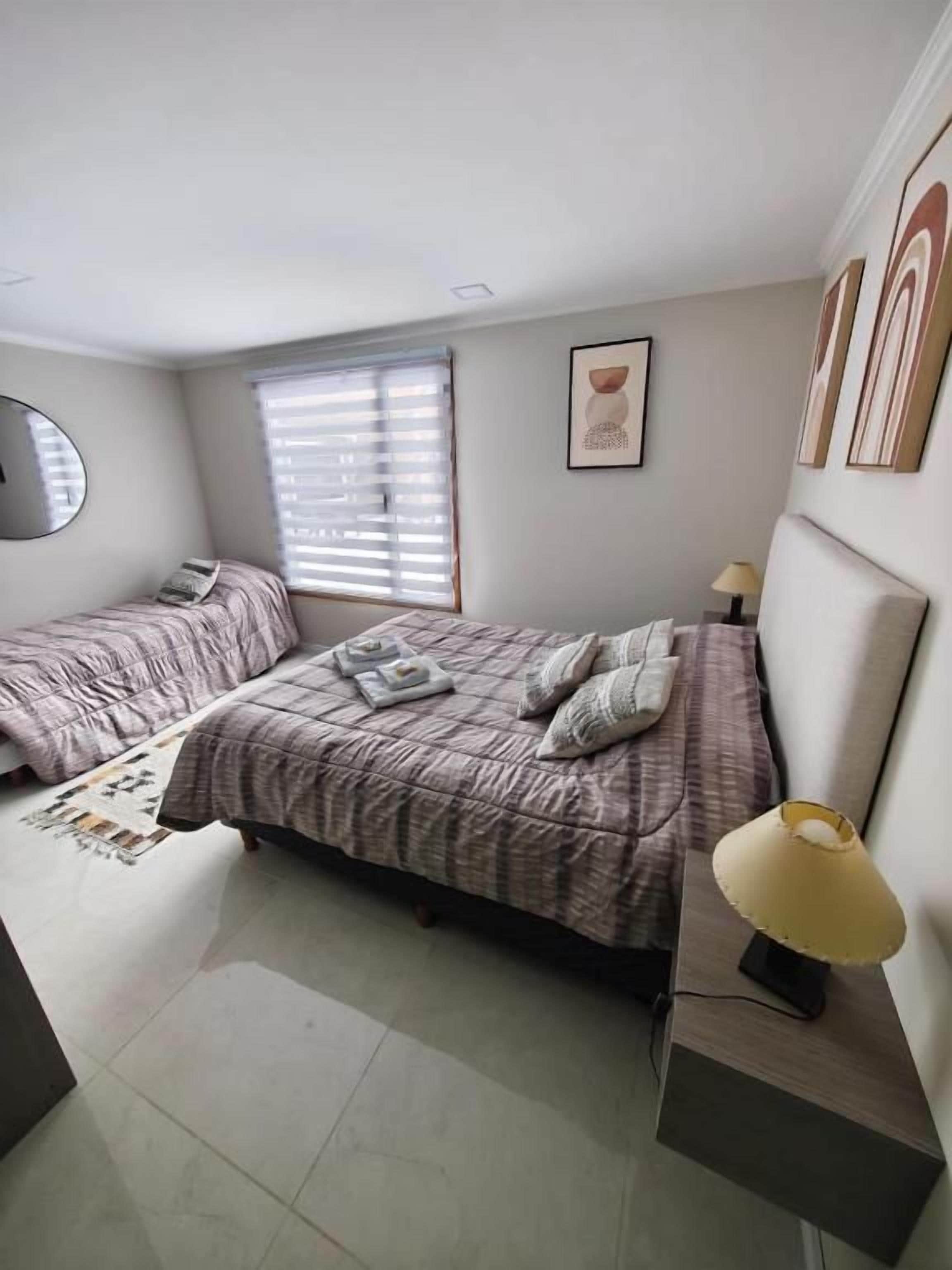 Un dormitorio con dos camas, una mesita de noche con lámpara, una ventana con persianas y un cuadro en la pared.