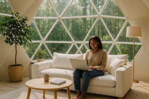 A person sitting on a white sofa with a laptop, a round wooden table with a book and a mug, and a potted plant beside the sofa. - Surf City Resorts (El Tunco)
