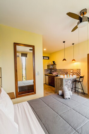 Una camera d'albergo con un letto, una zona salotto con tavolo e sedie, un angolo cottura e una ventola a soffitto.