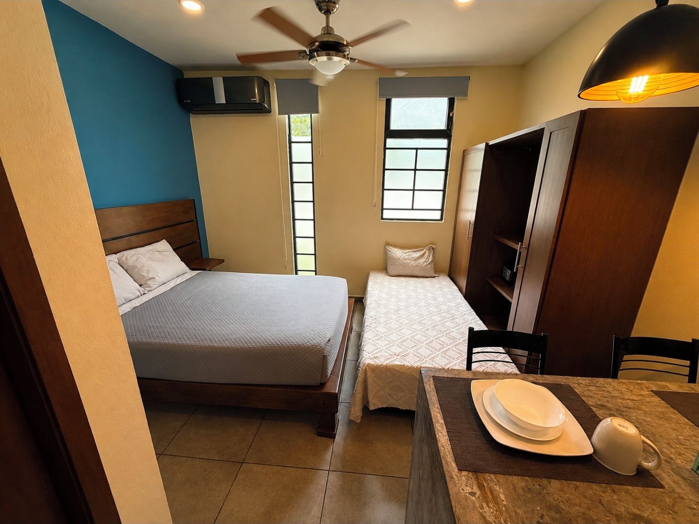 A bedroom with a bed, a smaller bed, a desk with a chair, a wardrobe, and a ceiling fan.