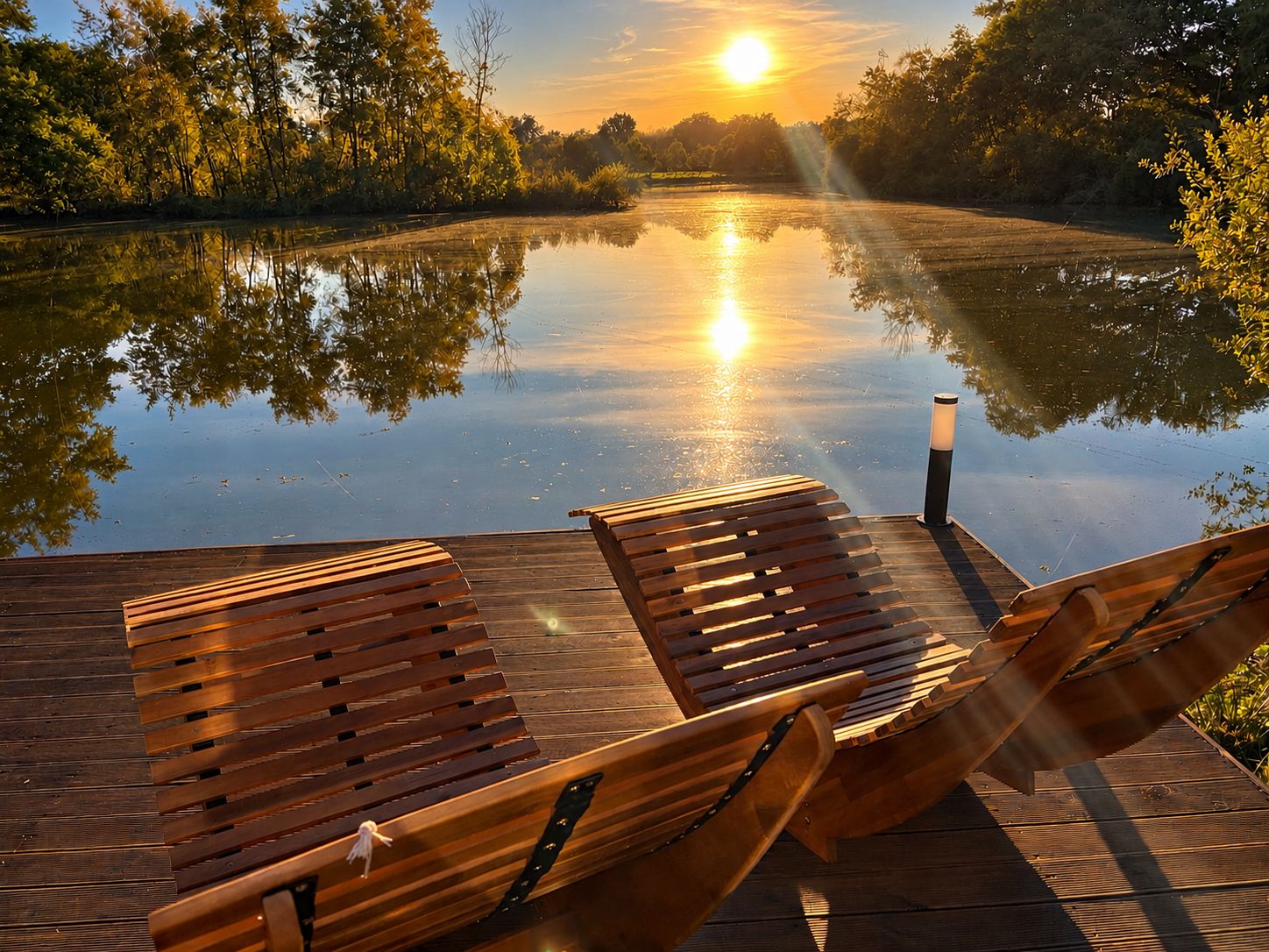 Een houten terras met twee loungestoelen gericht op het kalme wateroppervlak dat de zon weerkaatst.