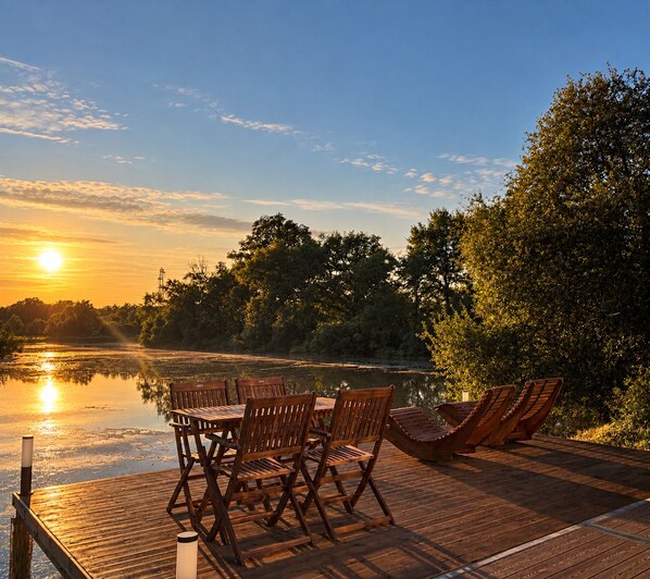 A wooden deck with a table and chairs overlooking a calm river at sunset.