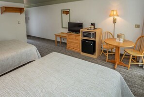 Ein Hotelzimmer mit zwei Betten, einem Schreibtisch mit Fernseher, einem Kühlschrank, einer Mikrowelle, einer Lampe und einem kleinen Tisch mit Stühlen.