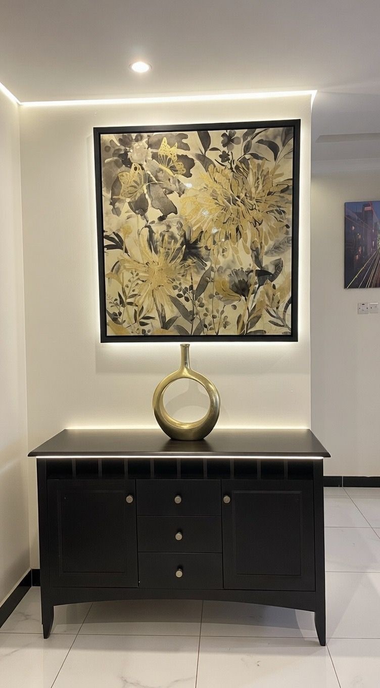 A black console table with a gold decorative object on top, a framed floral artwork above it, and a small framed picture on the wall to the right.