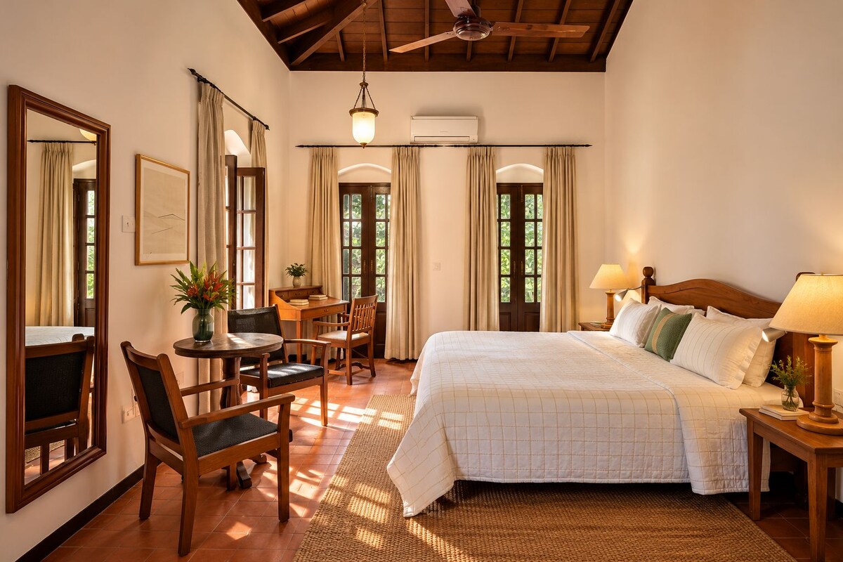 A hotel room with a bed, bedside tables, a desk with chairs, a ceiling fan, and large windows.