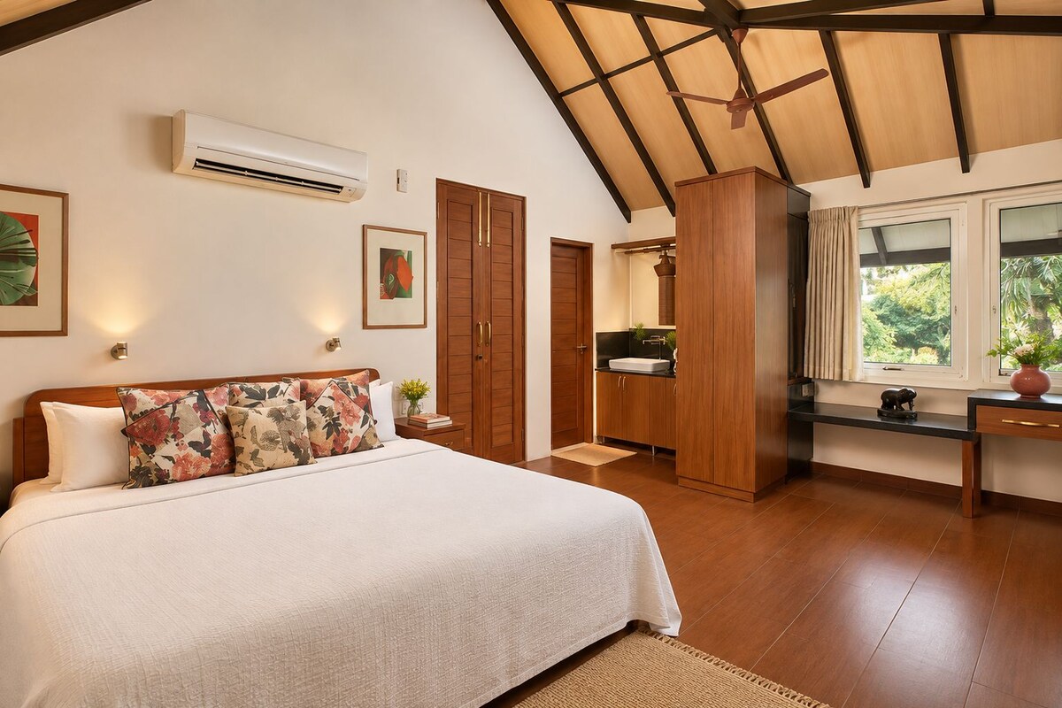 A bedroom with a large bed, a wardrobe, a desk, and a window with a view of greenery.