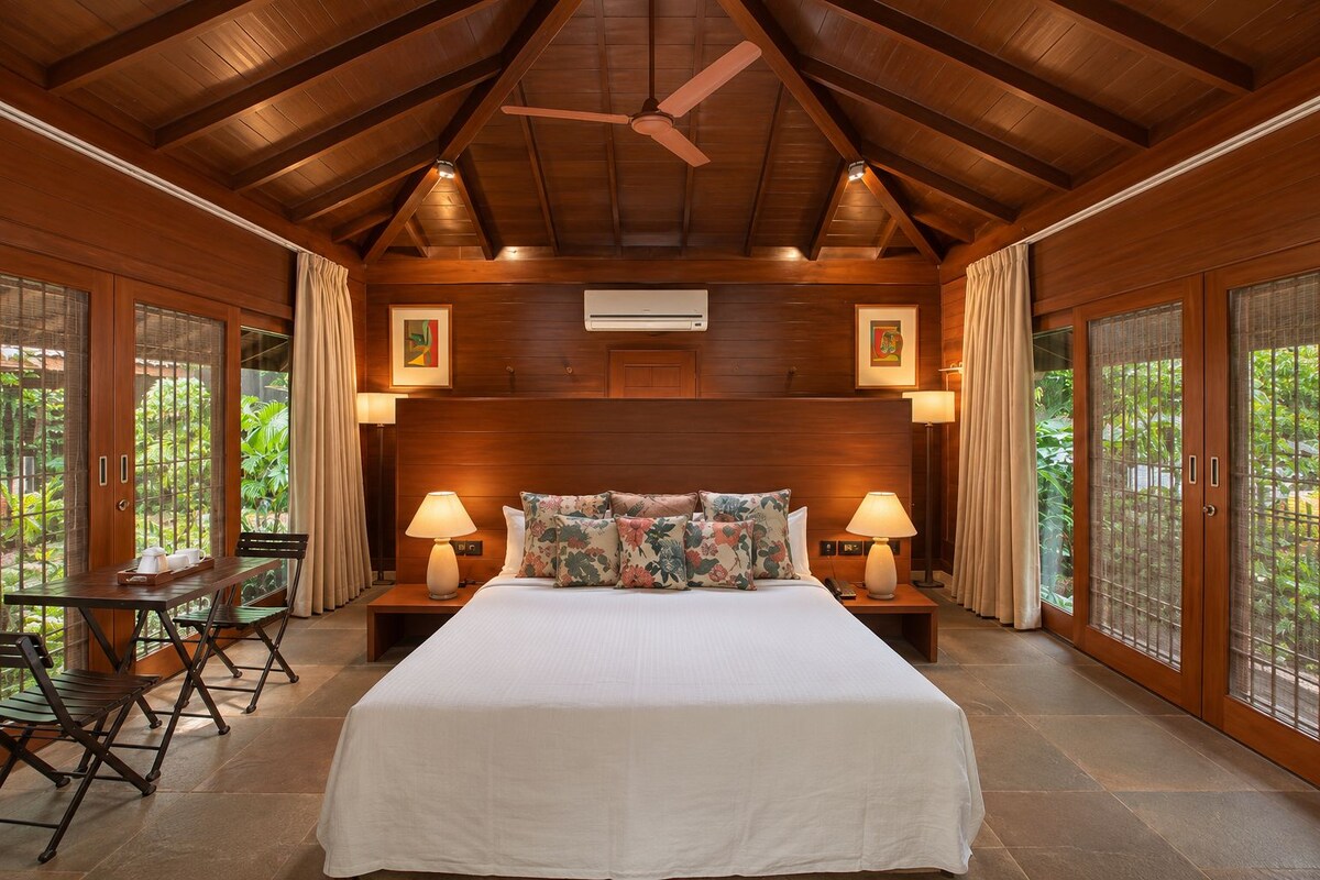 A spacious room with a large bed, wooden ceiling, and a view of greenery outside.