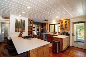 A modern kitchen with wooden cabinets, a central island, and a view of the outdoors through a glass door. - The Gallatin Riverstead (Big Sky)