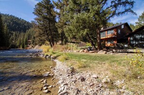 A cabin by a river with a clear view of the water and surrounding forest. - The Gallatin Riverstead (Big Sky)