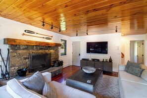 A cozy living room with a stone fireplace, wooden ceiling, and a flat-screen TV mounted on the wall. - The Gallatin Riverstead (Big Sky)