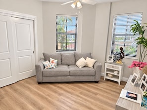 A living room with a grey sofa, two windows with blinds, a ceiling fan, and a small white side table with decorative items. - Cape Harbour Marina Condo with Huge Private Veranda (Cape Coral)