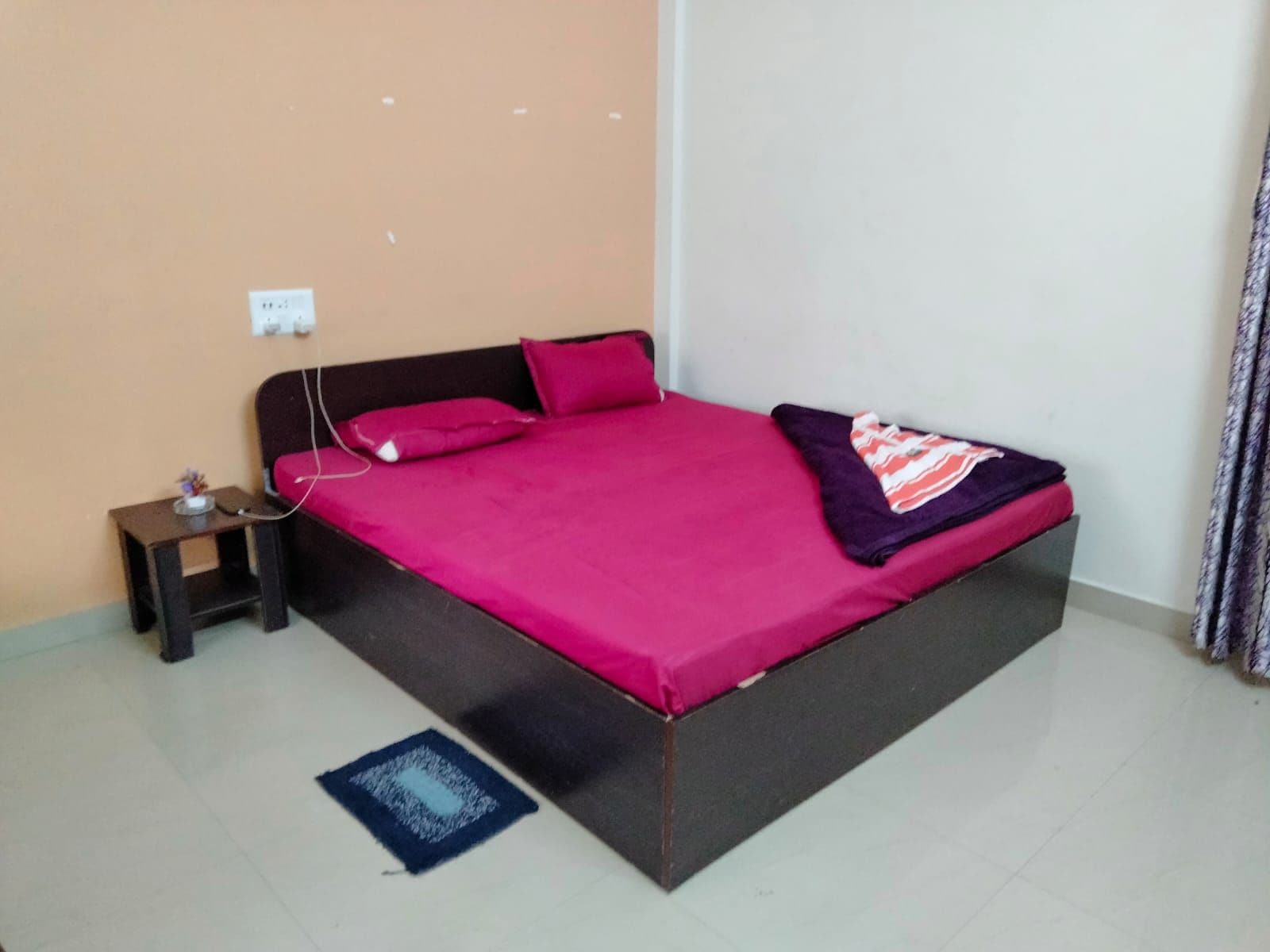A single-bed room with a dark headboard, a pink bedspread, a small bedside table with a lamp, and a power outlet on the wall.