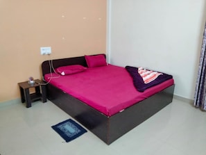 Quarto individual com uma cabeceira escura, uma colcha rosa, uma pequena mesa de cabeceira com abajur e uma tomada na parede.