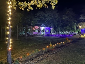 Área externa iluminada à noite por luzes de corda, com um pequeno edifício e um caminho.