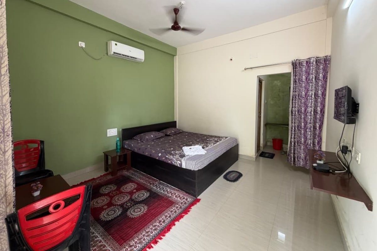 Quarto com cama, ventoinha no teto, ar condicionado, televisão, cadeira vermelha, mesa pequena e tapete estampado.