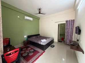 Quarto com cama, ventoinha no teto, ar condicionado, televisão, cadeira vermelha, mesa pequena e tapete estampado.